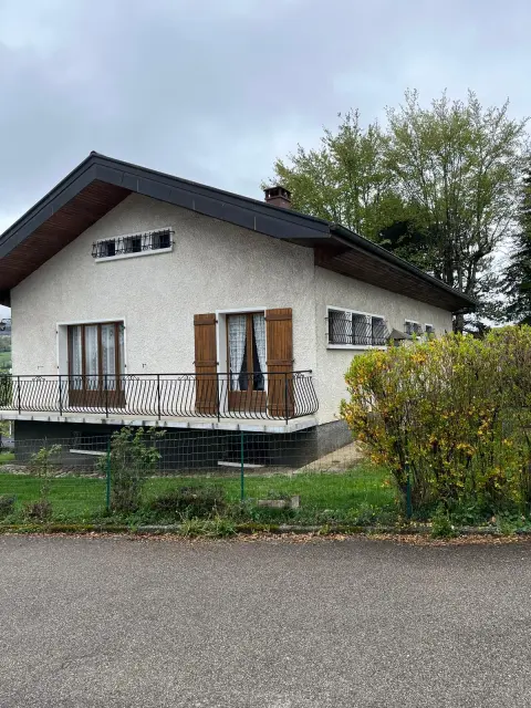 La maison et son terrain engazonné, volets de bois clair et toit de tuiles brunes, sous un ciel de printemps.
