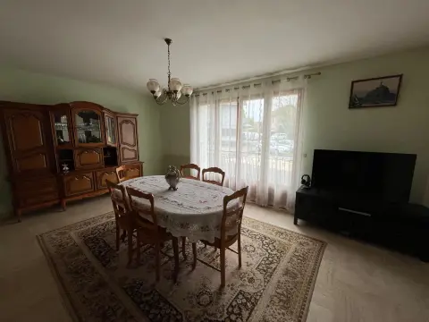 Le séjour avec la table, le tapis et le coin télévision, éclairé par les rideaux de la baie.